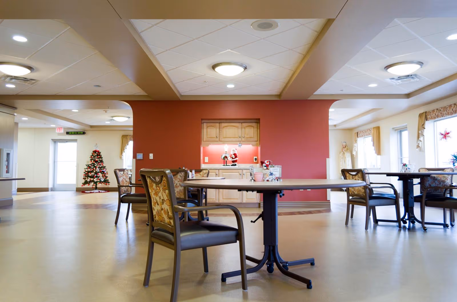 A spacious common area in a senior living facility with round tables and chairs arranged for seating. The room has a beige floor, a red accent wall with a small kitchenette or serving area featuring wooden cabinets and holiday decorations including a Santa figurine. There is a decorated Christmas tree near a door in the background, and large windows with curtains letting in natural light.