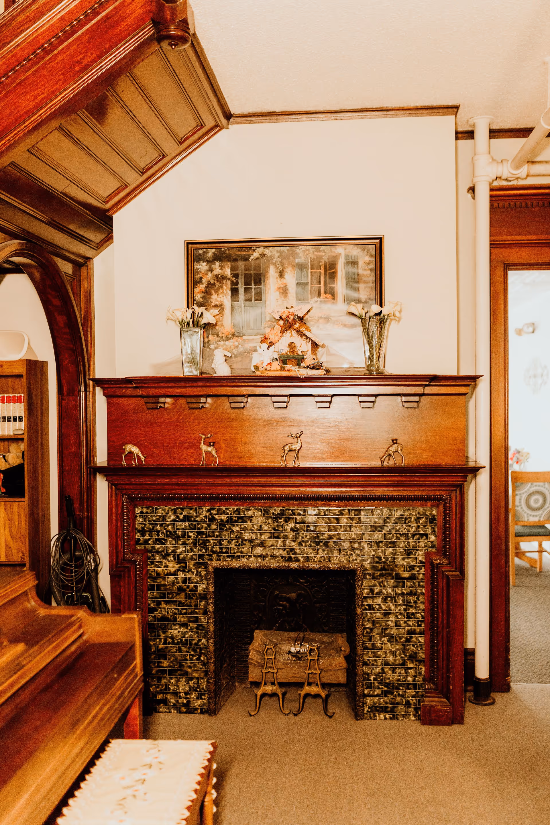 A cozy interior scene featuring a decorative fireplace with a dark tiled surround and a wooden mantel adorned with small deer figurines, vases with flowers, and a framed painting above. To the left, part of a wooden piano and bookshelf with books are visible. The room has warm wood accents and beige carpeting.