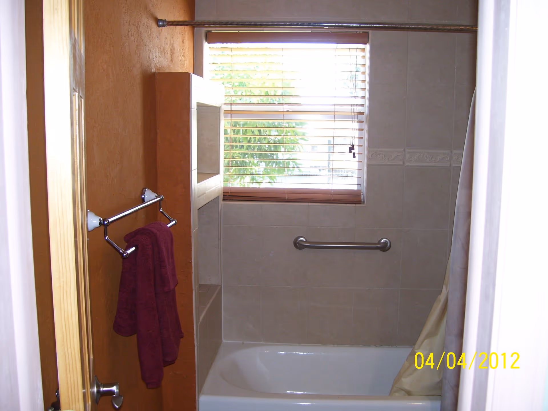 Bathroom with a bathtub and shower combination. The walls are tiled in beige with a decorative border. There is a window with wooden blinds above the bathtub. A metal grab bar is installed on the wall inside the bathtub area. A towel rack with a maroon towel is mounted on the left wall, which is painted a warm brown color. A shower curtain is partially visible on the right side.