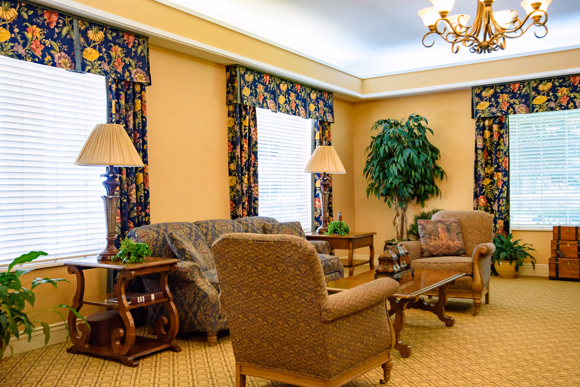 A cozy living room with patterned armchairs and a sofa arranged around a glass coffee table. The room has large windows with floral curtains and valances, two table lamps on wooden side tables, a potted plant in the corner, and a chandelier hanging from the ceiling. The carpet has a geometric pattern.