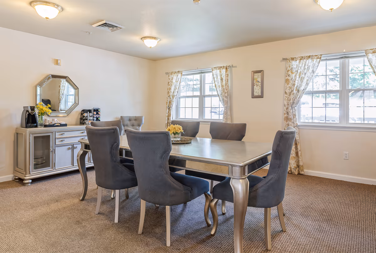 A dining room with a rectangular table surrounded by six gray upholstered chairs. The room has beige walls, two windows with floral curtains, a sideboard with a mirror above it, and a coffee maker on top. The floor is carpeted and the room is well-lit with ceiling lights.