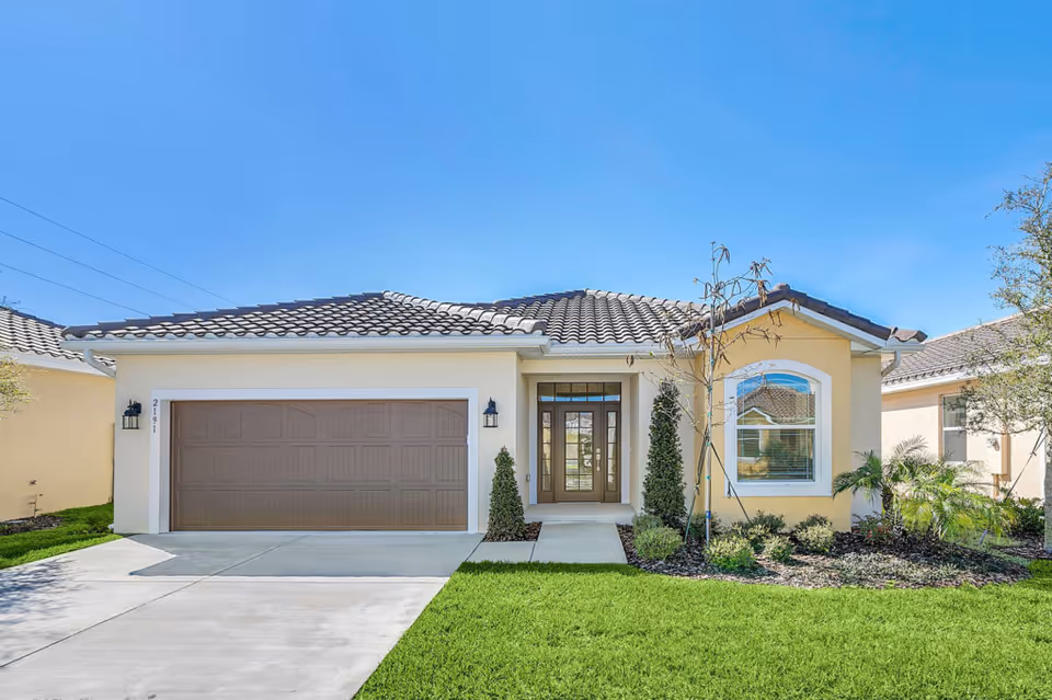 Front exterior view of a single-story house with a tiled roof, a double garage door, a walkway leading to a glass-paneled front door, and a landscaped yard with green grass, small trees, and shrubs under a clear blue sky.