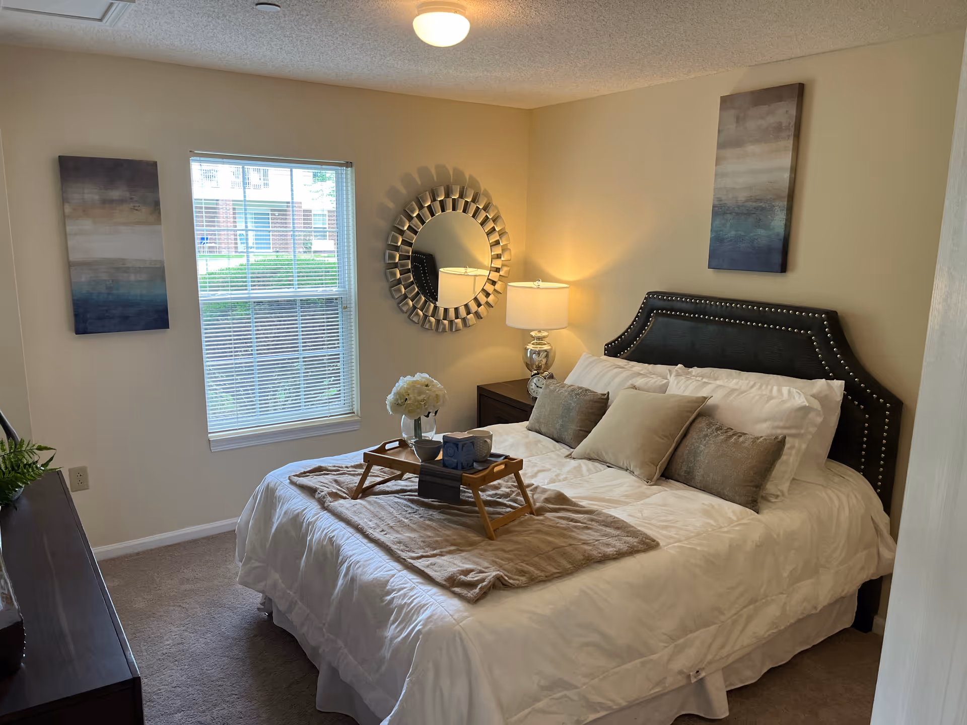 A neatly made bed with white bedding and multiple pillows in a bedroom. A wooden tray with a vase of white flowers and two mugs is placed on the bed. The room has beige walls with two abstract paintings and a decorative round mirror above a nightstand with a lamp. A window with blinds lets in natural light, and a dresser with a plant is visible on the left side.