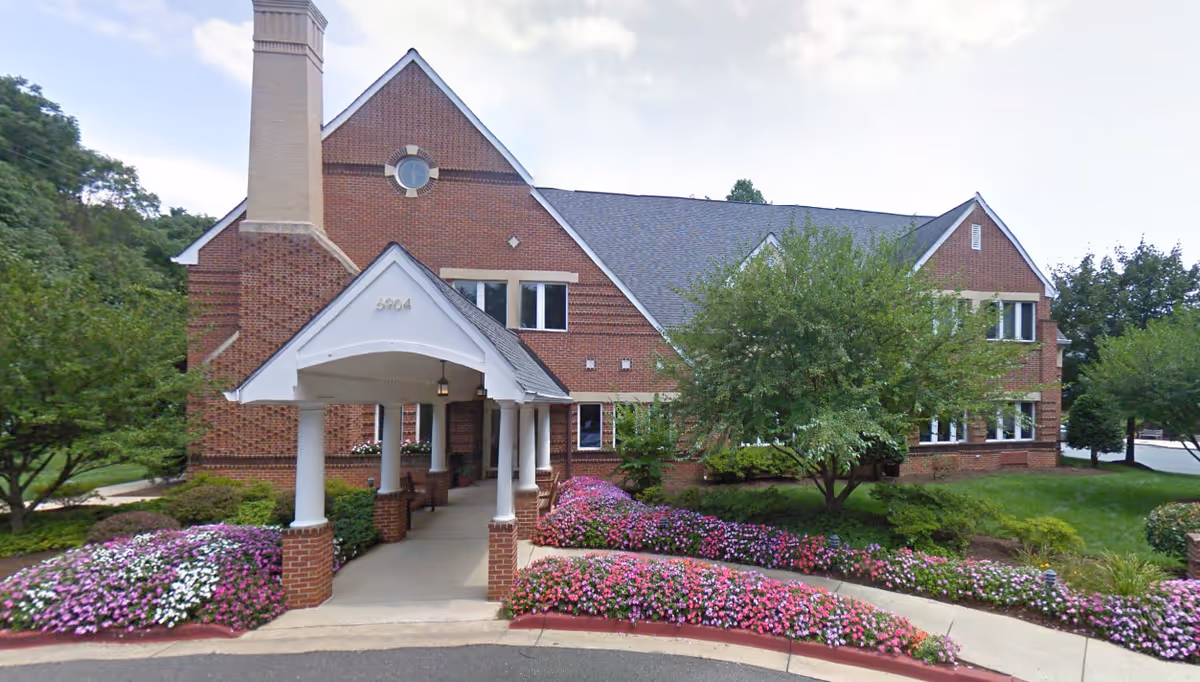 Exterior view of a two-story brick building with a covered entrance supported by white columns. The building is surrounded by well-maintained landscaping including green trees, bushes, and colorful flower beds along the walkway and driveway.