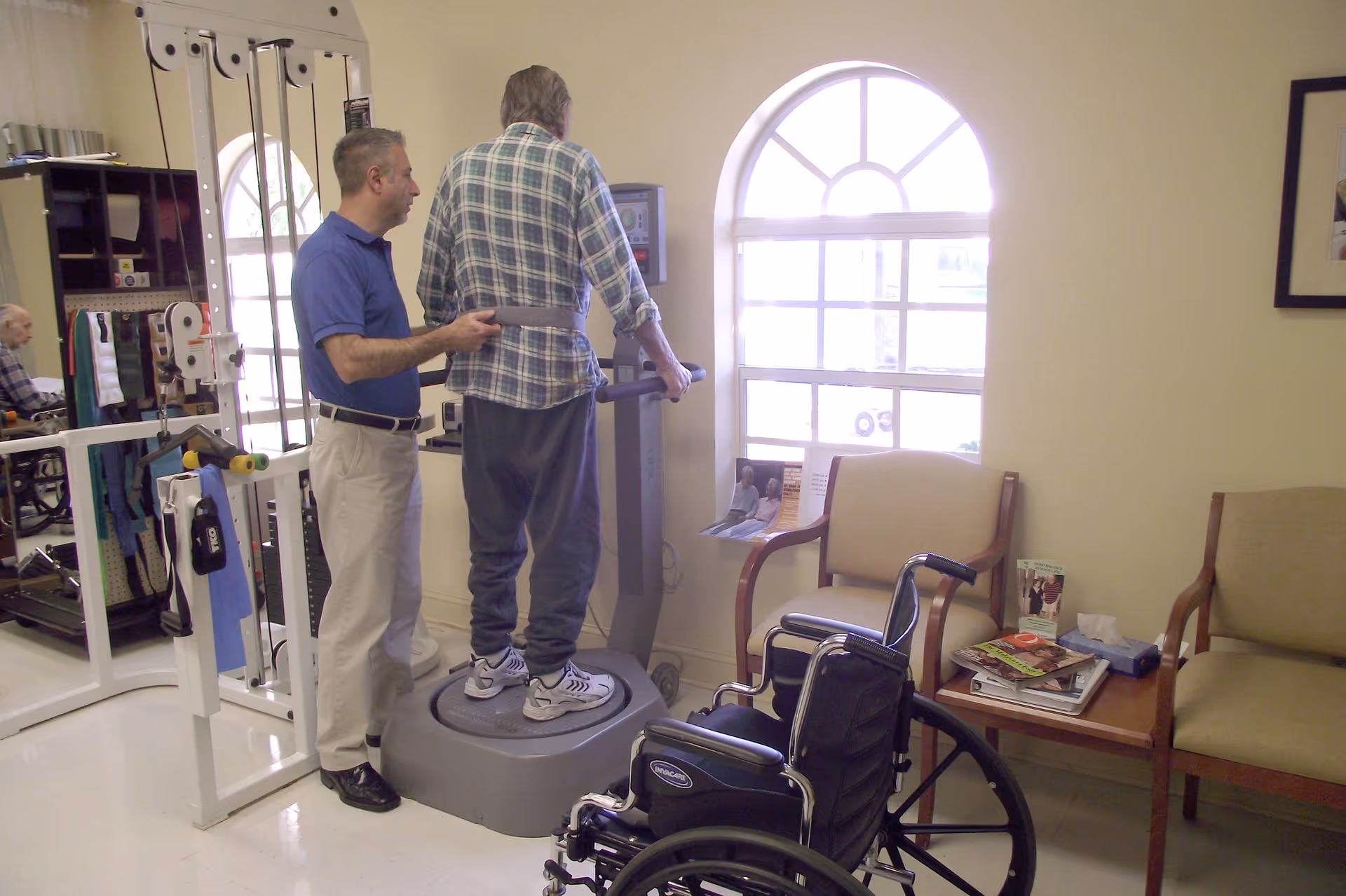 A man in a blue shirt assists an elderly man standing on a vibration exercise platform in a rehabilitation center room. There is a wheelchair in the foreground, chairs and a small table with magazines near a window with an arched top. Exercise equipment and another elderly man in a wheelchair are visible in the background.