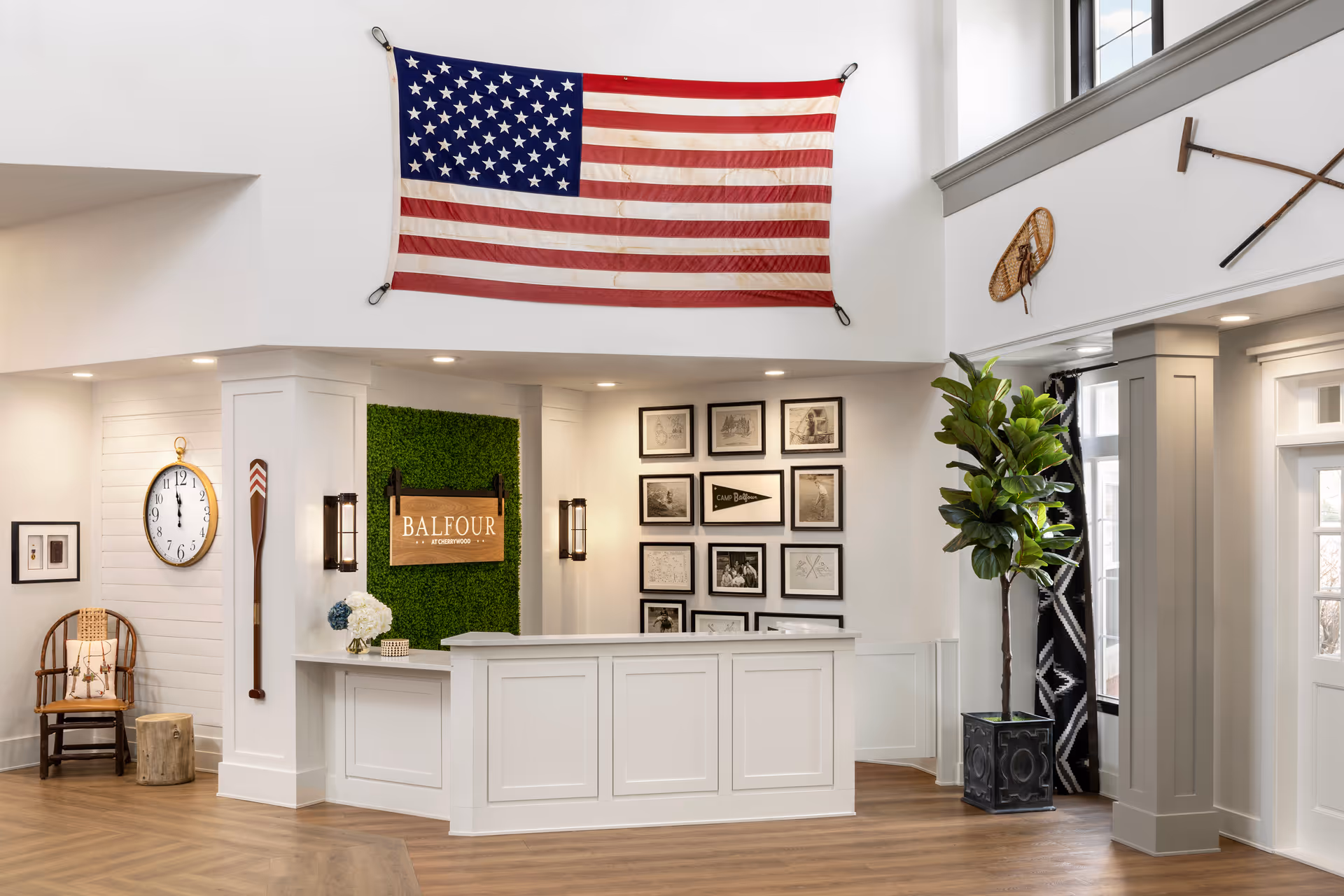 Bright reception lobby with a white front desk, a 'Balfour' sign on a green wall, framed photos, an American flag overhead, and seating.