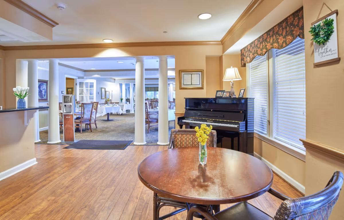 Bright communal dining/lounge area with a round table and vase of yellow flowers, a piano by the window, and columns opening into a dining room.