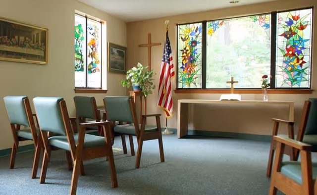 A small chapel room with wooden chairs arranged in rows facing an altar. The altar has a cross, an open book, and a vase with a single red flower. Behind the altar are large stained glass windows with colorful floral designs. An American flag and a wooden cross are positioned near the windows. The room has beige walls and carpeted floor.