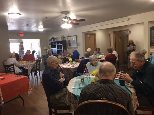 A group of elderly people sitting at round tables covered with floral tablecloths in a dining room. They are engaged in conversation and enjoying drinks. The room has wooden floors, ceiling fans with lights, and shelves with decorations in the background.
