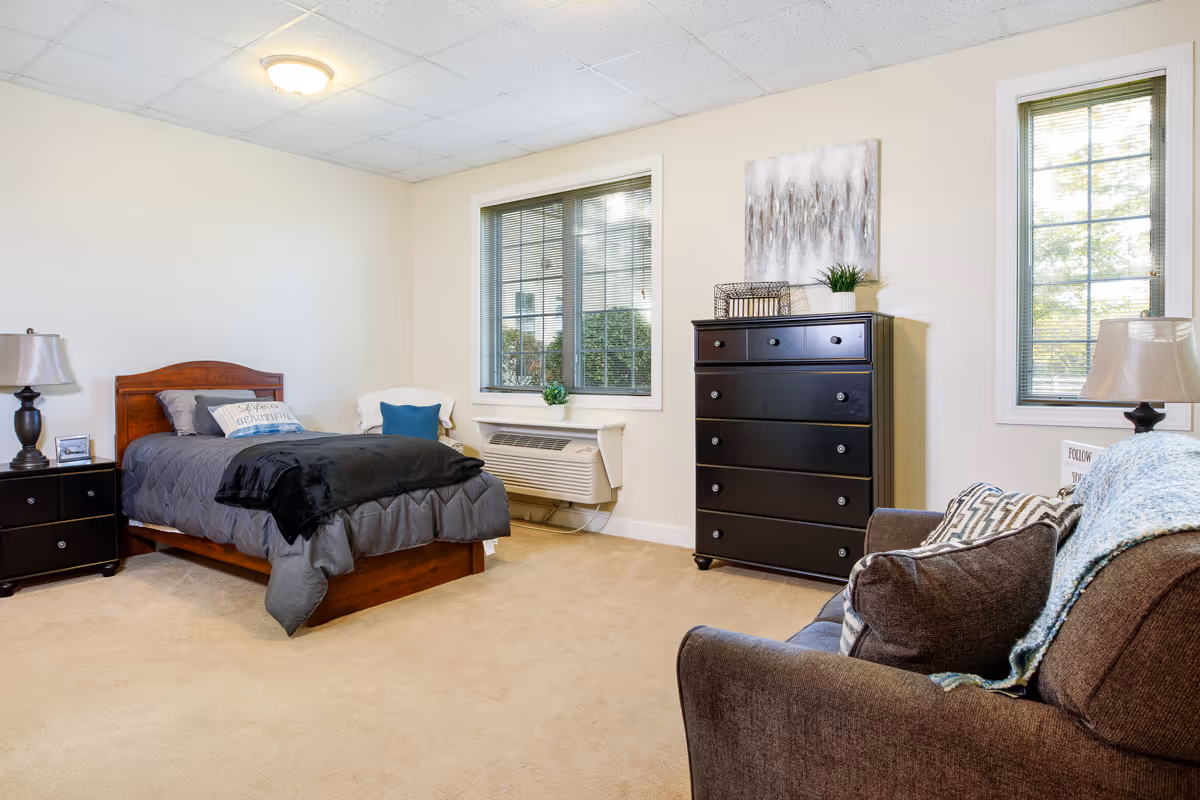 A cozy bedroom in a senior living facility featuring a single wooden bed with gray bedding and a decorative pillow, a dark wooden nightstand with a lamp and picture frame, a dark wooden dresser with decorative items and a painting above it, two windows with blinds, a brown upholstered armchair with cushions and a throw blanket, and beige carpeted floor.