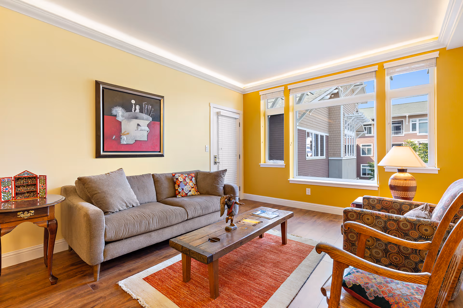 A cozy living room with yellow walls and large windows letting in natural light. The room features a gray sofa with colorful pillows, a wooden coffee table on a red and white rug, two patterned armchairs, a side table with a decorative item, and a table lamp. Outside the windows, parts of neighboring buildings are visible.
