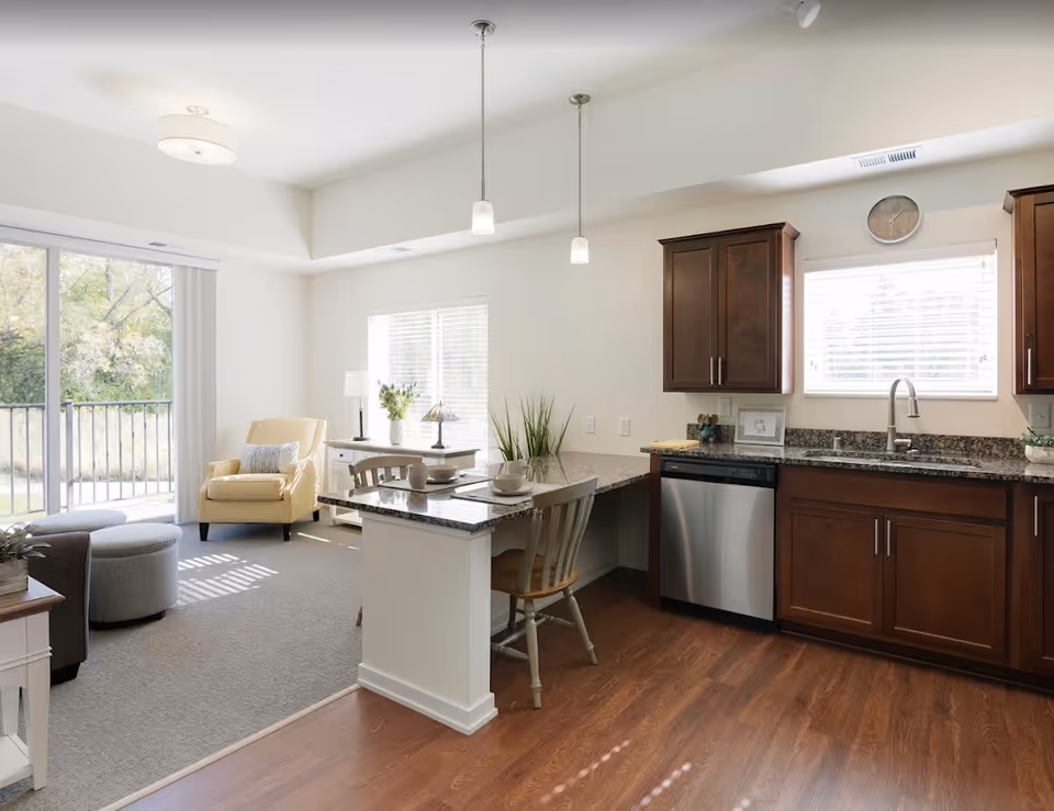 Bright and airy senior living apartment interior featuring a kitchen with dark wood cabinets, granite countertops, a stainless steel dishwasher, and a sink under a window with blinds. Adjacent to the kitchen is a small dining area with a table set for two and a wooden chair. The living area includes a yellow armchair, a gray ottoman, a small side table with a lamp, and large sliding glass doors leading to an outdoor balcony with a view of trees.