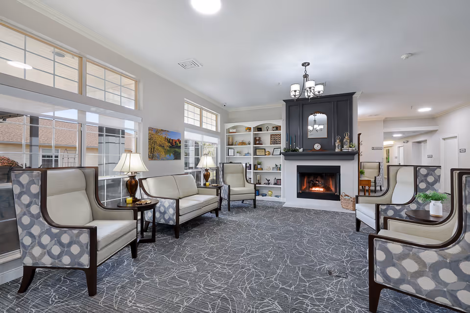A bright and spacious living room in an assisted living facility featuring several cushioned armchairs and a loveseat arranged around a fireplace with a lit fire. The room has large windows allowing natural light to fill the space, a patterned carpet, two table lamps on side tables, and built-in shelves with decorative items and books.