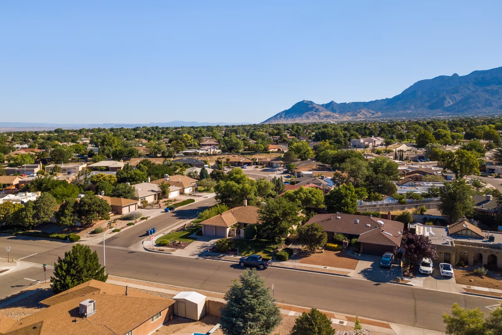 Aerial view of a suburban neighborhood with single-story houses, streets, and trees, with mountains visible in the background under a clear blue sky.