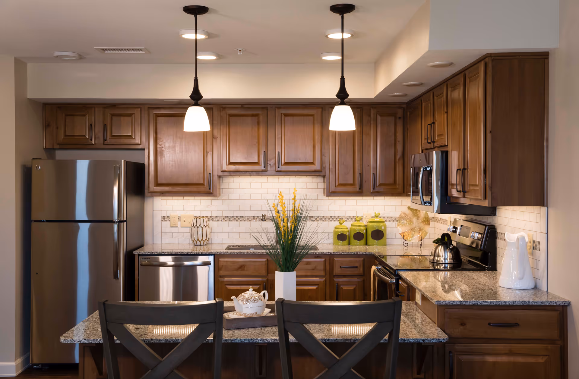 Modern kitchen with wooden cabinets, stainless steel refrigerator, dishwasher, microwave, and stove. Granite countertops with a small island featuring two chairs, a white teapot on a tray, and a vase with yellow flowers. Two pendant lights hang from the ceiling.