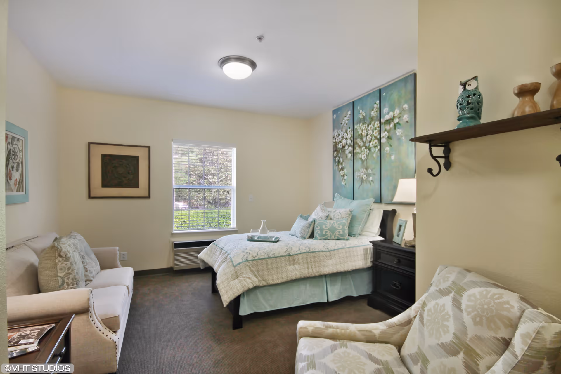 A cozy bedroom with a bed adorned with light blue and white bedding, a small sofa with patterned cushions, an armchair, and a window letting in natural light. The walls are decorated with framed artwork and a shelf holding decorative items.