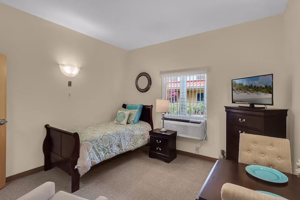 Furnished bedroom with a single bed, nightstand and lamp, dresser with TV, window air conditioner and a small table and chairs.