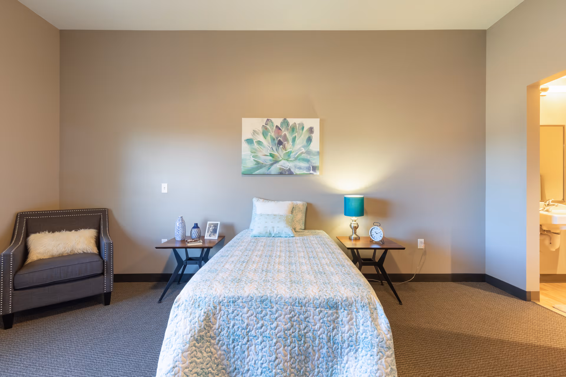 A neatly made single bed with bedside tables, a lamp, an armchair, and wall art in a neutral-toned bedroom.
