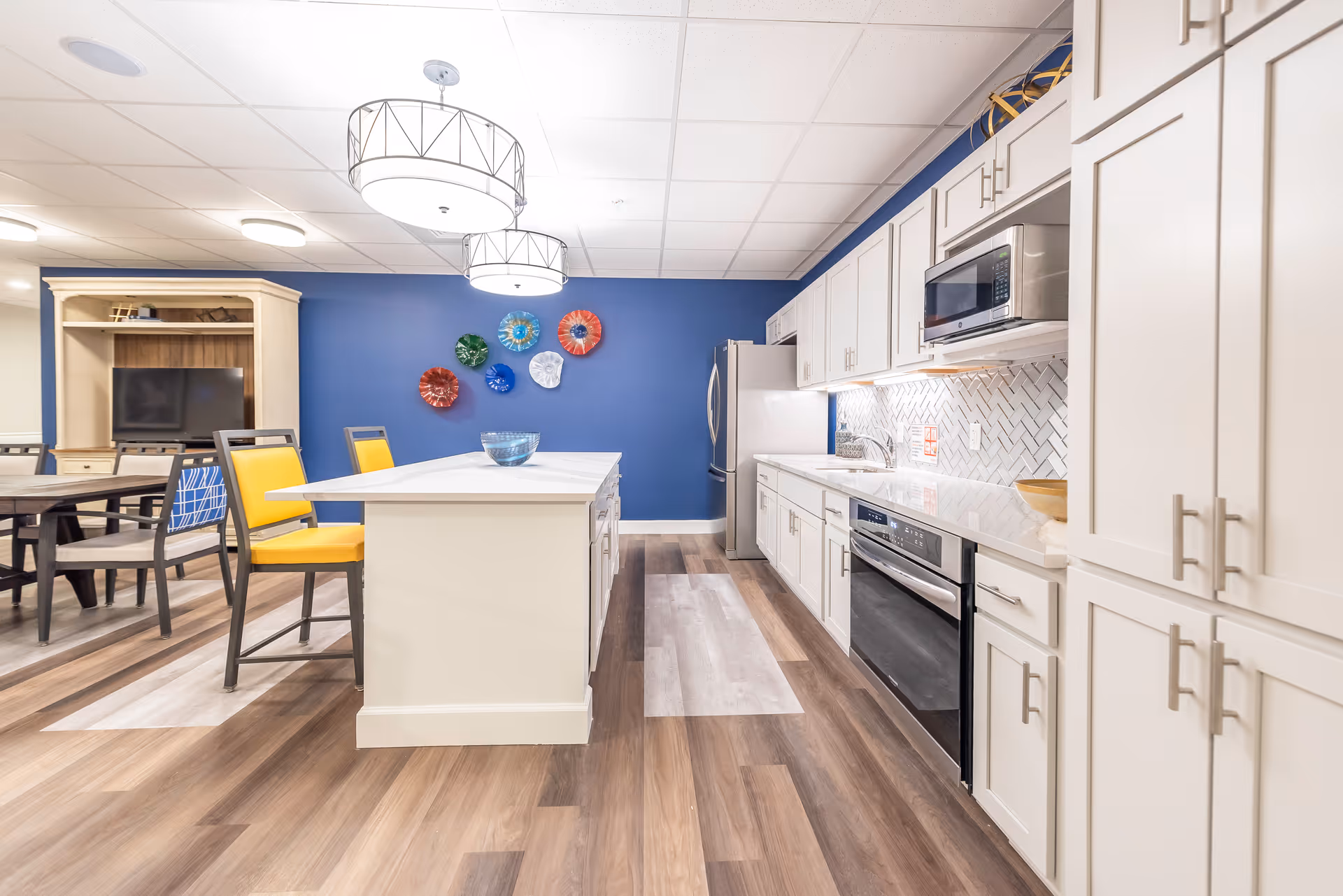A modern kitchen area with white cabinetry, stainless steel appliances including a microwave and oven, and a large island with yellow chairs. The floor has a wood pattern, and the back wall is painted blue with colorful decorative plates mounted on it. In the background, there is a dining area with tables and chairs and a TV on a wooden stand.