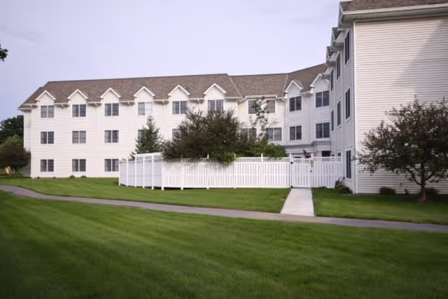 Exterior view of a multi-story senior living facility building with white siding, multiple windows, and a white picket fence enclosing a small garden area. There is a paved walkway and well-maintained green lawn in the foreground under a partly cloudy sky.