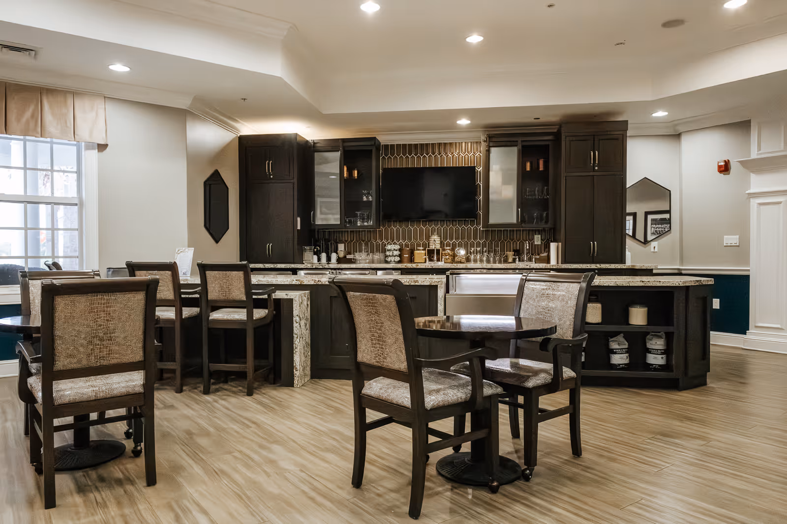 A modern kitchen and dining area with dark wood cabinets, a granite countertop island, and several tables with cushioned chairs. The kitchen features a tiled backsplash, a mounted flat-screen TV, and various kitchen items on the counter. The room has wood flooring and recessed lighting.