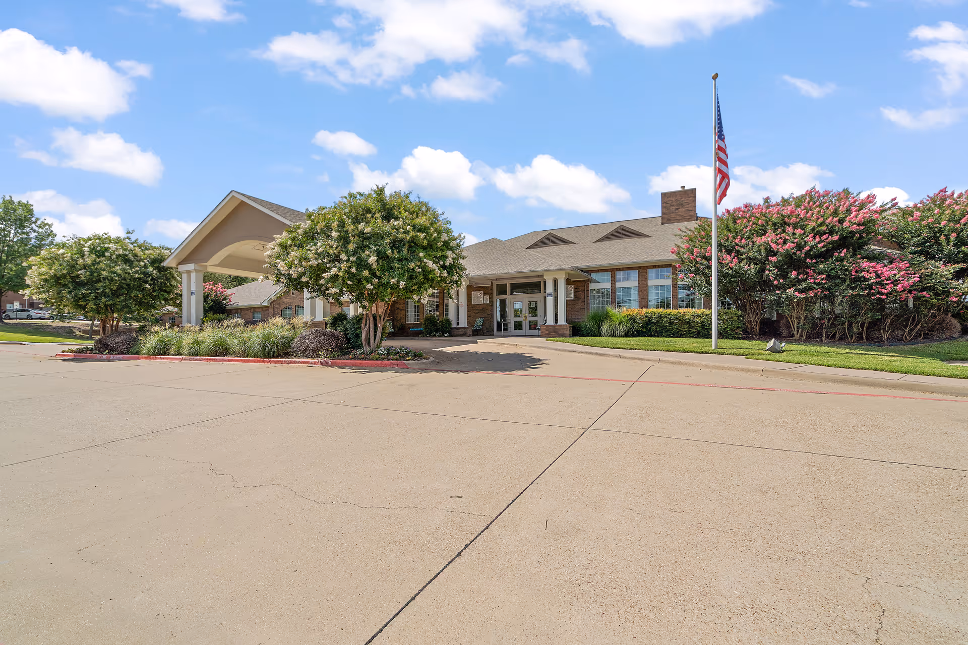 Front entrance of a single-story senior living facility with a covered porte-cochere, landscaped shrubs, and an American flag.