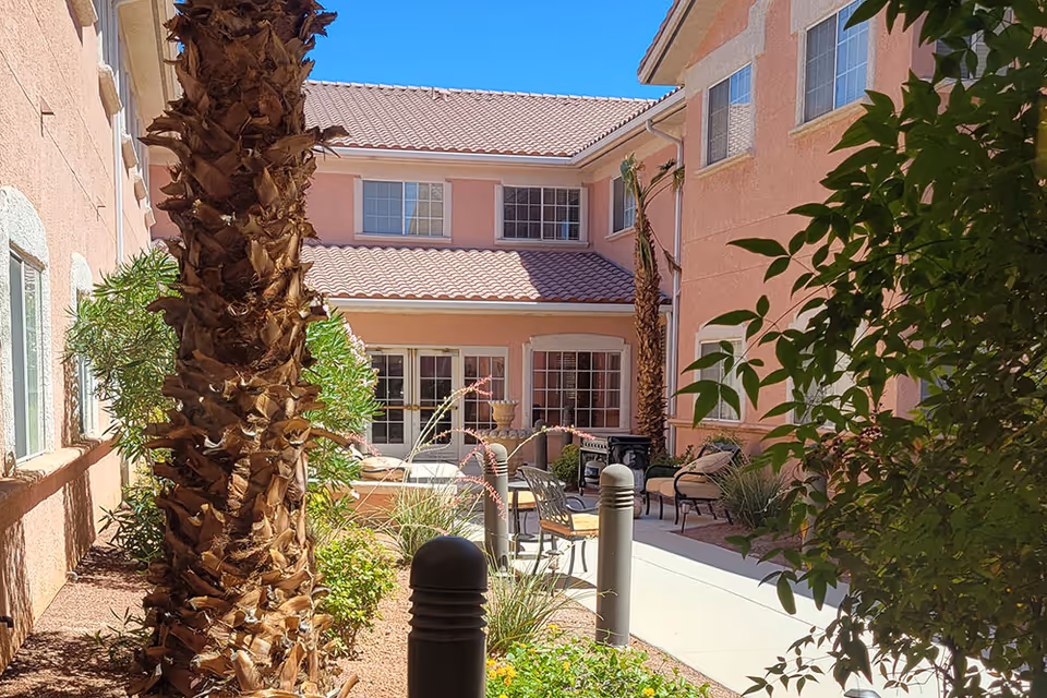 Outdoor courtyard area at The Bridge at Paradise Valley featuring a paved walkway, palm trees, various plants, and outdoor seating with chairs and a small table, surrounded by a two-story building with pink stucco walls and tiled roof under a clear blue sky.