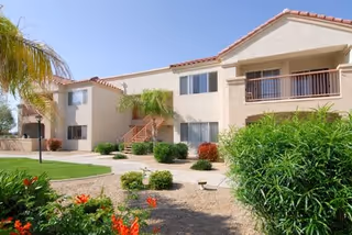 Exterior view of a two-story residential building with beige walls and red-tiled roof, surrounded by green shrubs, palm trees, and a landscaped garden with gravel and flowering plants under a clear blue sky.