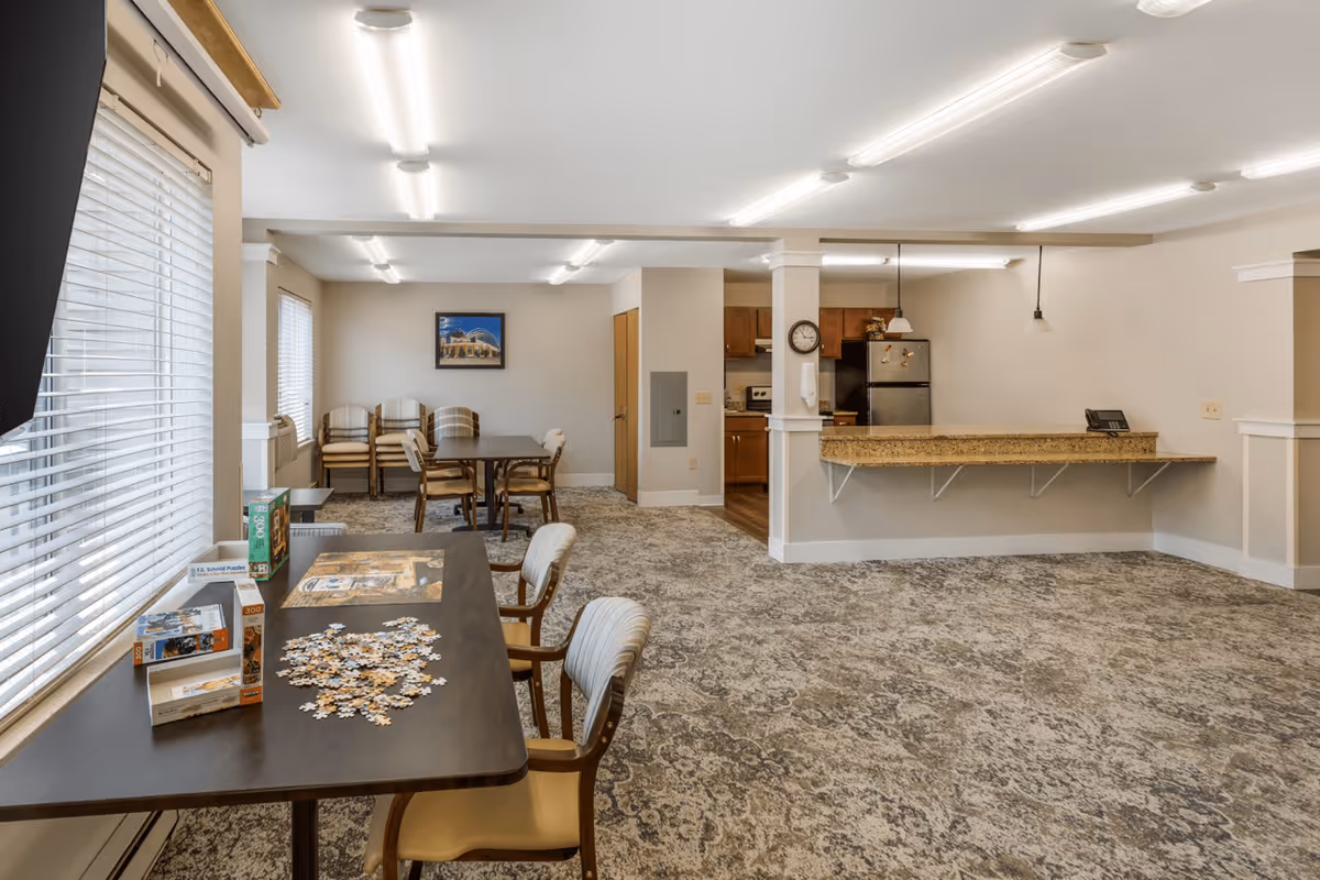 A spacious senior living common area with carpeted floors and beige walls. There are tables and chairs arranged for activities, including a table with a partially completed jigsaw puzzle near a window with blinds. In the background, there is a kitchen area with wooden cabinets, a refrigerator, and a granite countertop bar with hanging pendant lights. A clock is mounted on a column near the kitchen.