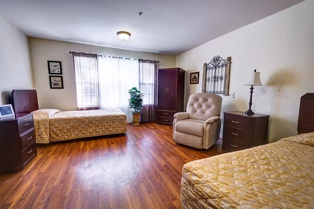 A bright bedroom with two single beds covered in beige quilted bedspreads, a beige recliner chair, two dark wooden nightstands with a lamp on one, a dark wooden wardrobe, framed artwork on the walls, a potted plant near a window with sheer curtains, and wood flooring.