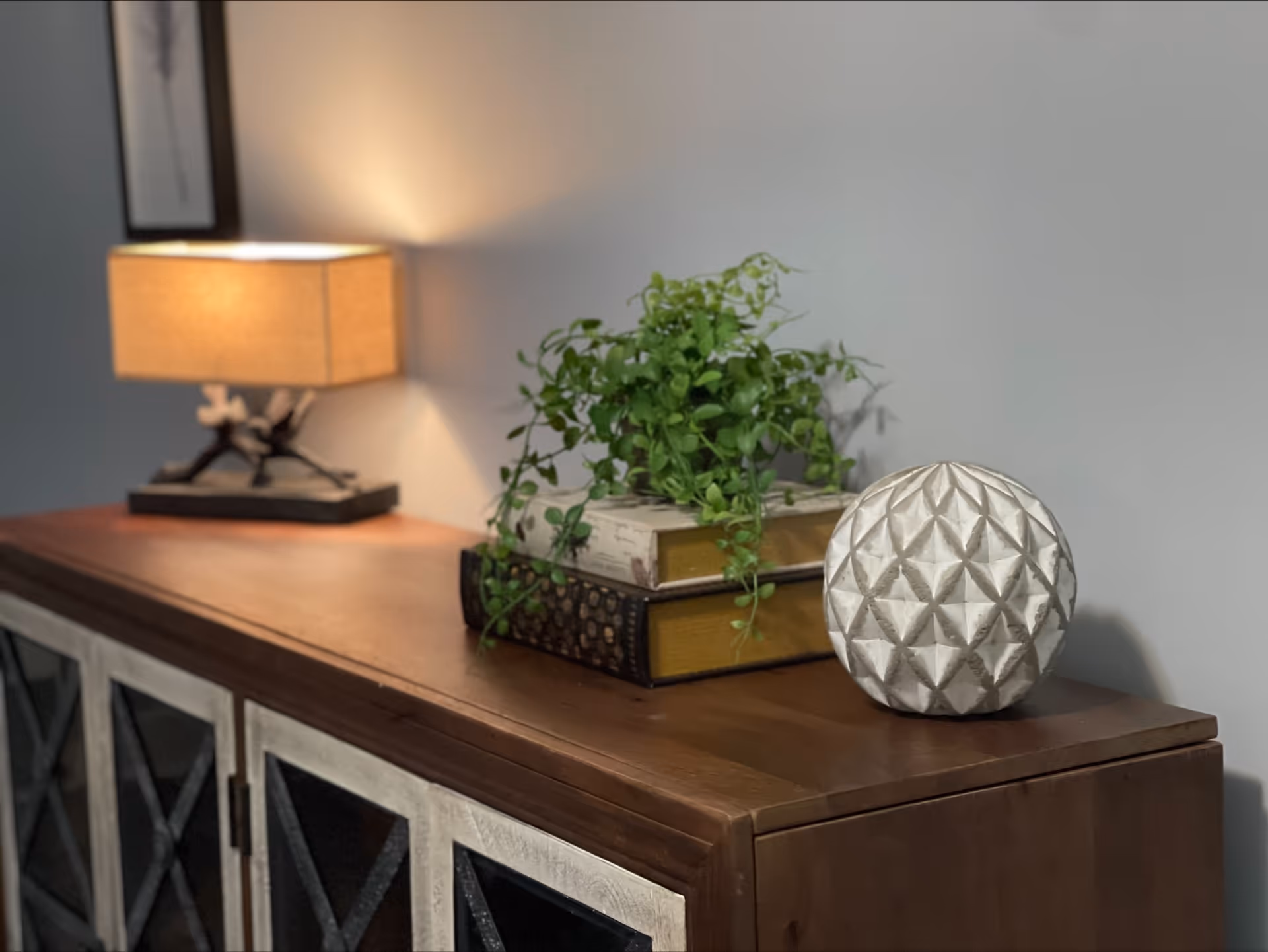 A wooden cabinet with a decorative table lamp, a green potted plant placed on two stacked books, and a white textured spherical ornament on top. The background wall is plain and light-colored.