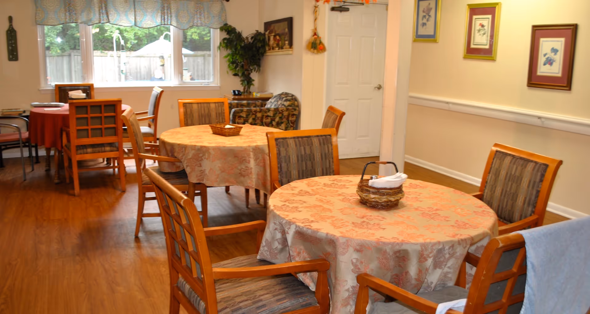 A cozy dining area with round tables covered in floral tablecloths and wooden chairs with cushioned seats. The room has wooden flooring, framed artwork on the walls, a window with a valance, and a door in the background. There is a small basket with napkins on the nearest table.
