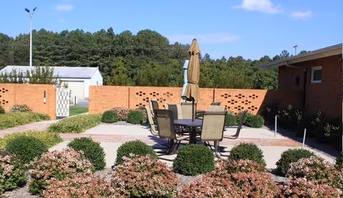 Outdoor courtyard patio with a round table, chairs and a closed umbrella surrounded by landscaping, a low brick wall, and buildings with trees in the background.