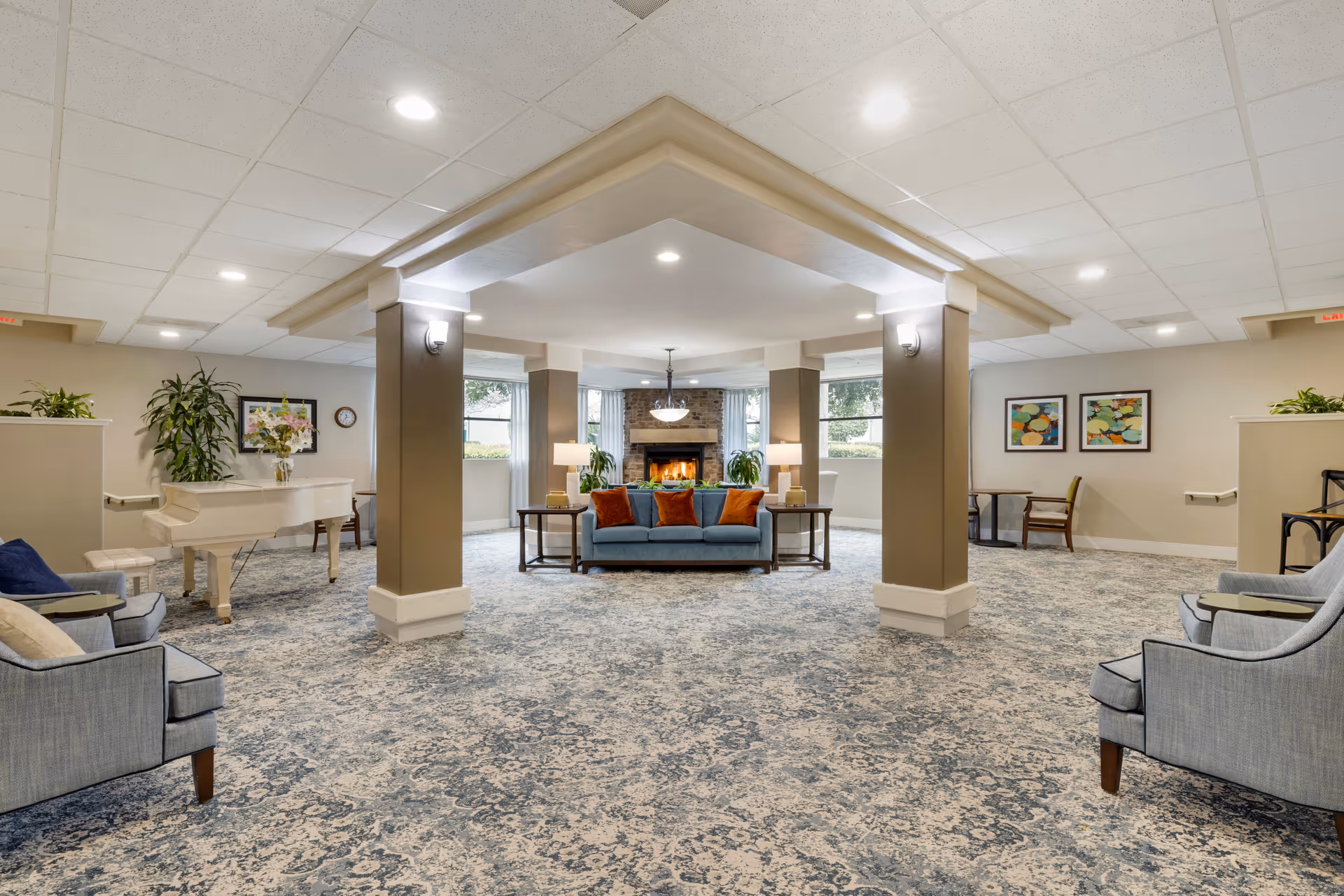 Spacious senior living common area with a blue sofa adorned with orange pillows centered in front of a stone fireplace. The room features beige walls, multiple windows with curtains, blue patterned carpet, several armchairs, a white grand piano with a vase of flowers, side tables with lamps, and decorative plants and artwork on the walls.