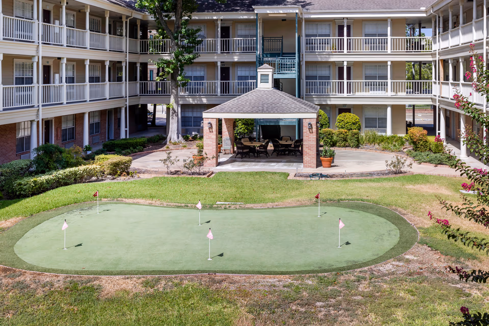 Outdoor courtyard area of a senior living facility with a putting green in the foreground, a covered seating area with chairs and tables in the middle, and a three-story building with balconies surrounding the courtyard.