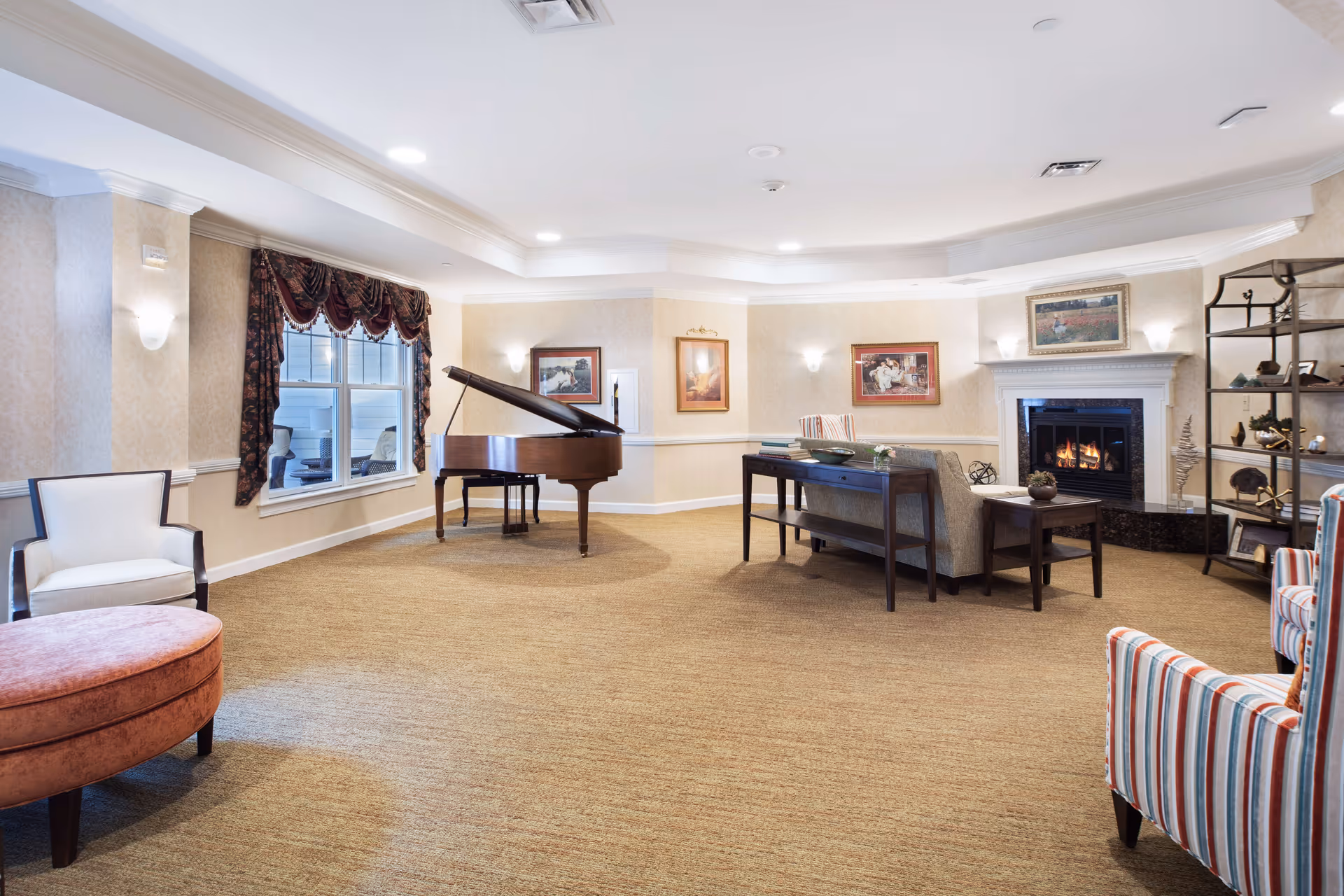 A spacious, well-lit living room with beige carpet and wallpapered walls. The room features a grand piano near a window with dark drapes, a fireplace with a fire burning, several framed paintings on the walls, a gray sofa with a wooden console table behind it, a striped armchair, a white armchair, a round pink ottoman, and a metal shelving unit with decorative items.