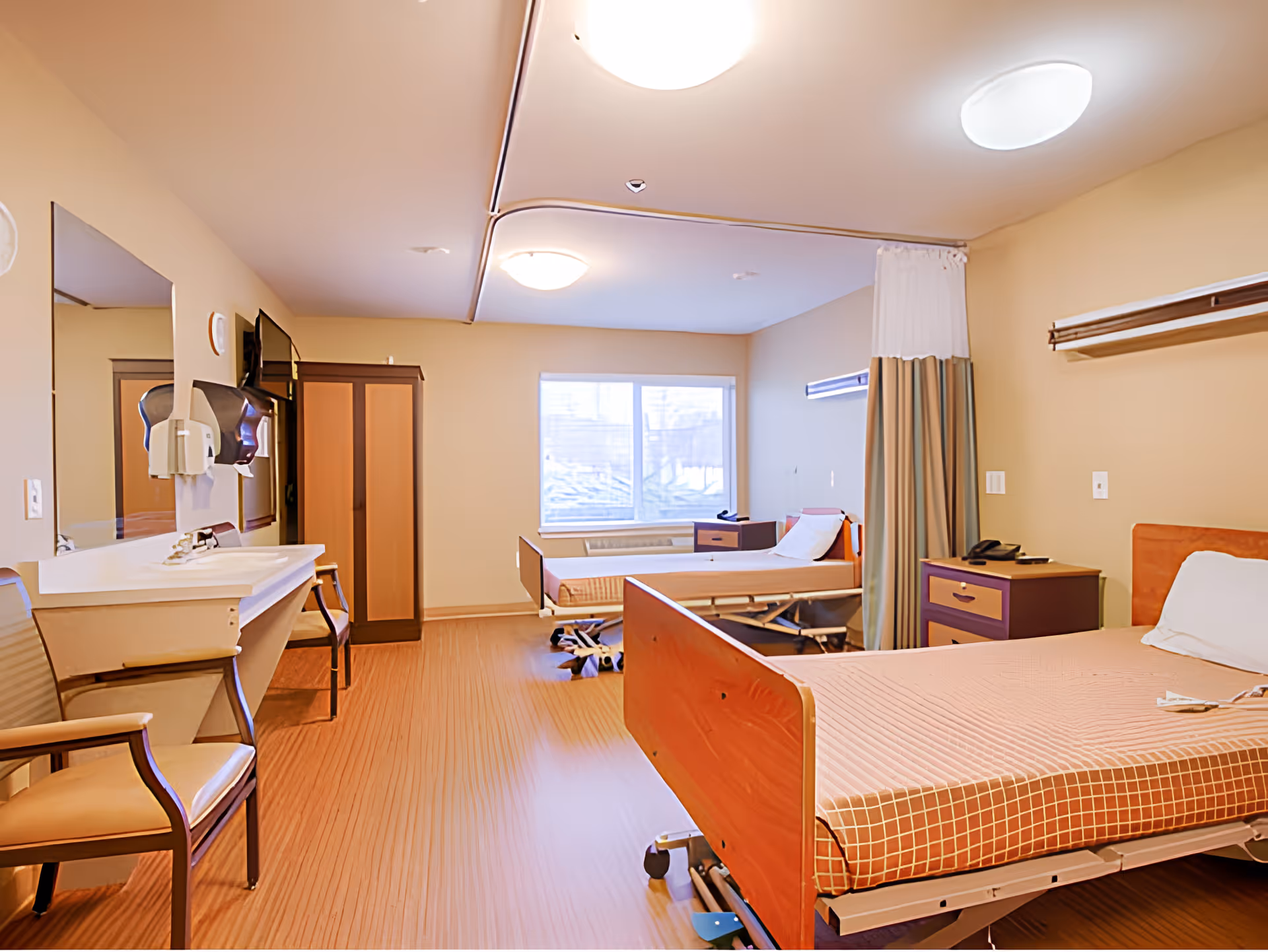 Bright shared resident bedroom with two adjustable hospital beds, bedside tables, chairs, a sink area and a window.