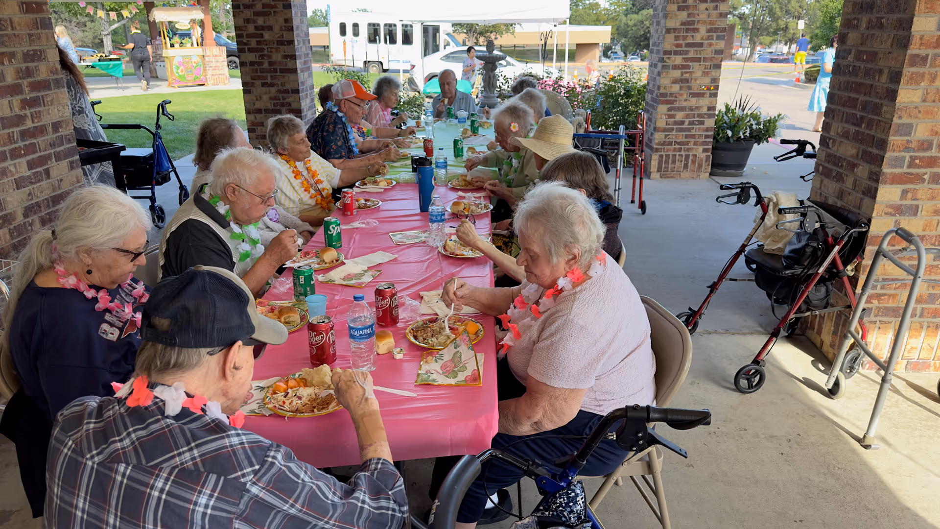 A group of elderly people sitting around a long table covered with a pink tablecloth, enjoying a meal together outdoors under a covered patio. Some are wearing colorful leis, and there are various drinks and plates of food on the table. Walkers and mobility aids are visible nearby, and a bus and other people can be seen in the background.