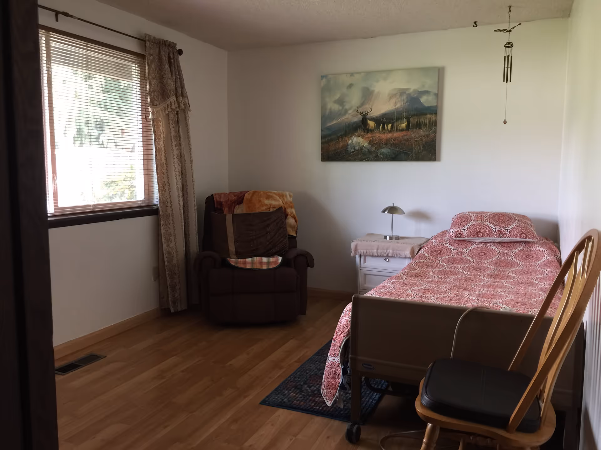 A simple bedroom with a single bed covered in a red patterned bedspread, a wooden chair with a black cushion, a small white nightstand with a lamp, a brown recliner chair with a blanket, a window with blinds and decorative curtains, and a painting of elk in a mountainous landscape on the wall.
