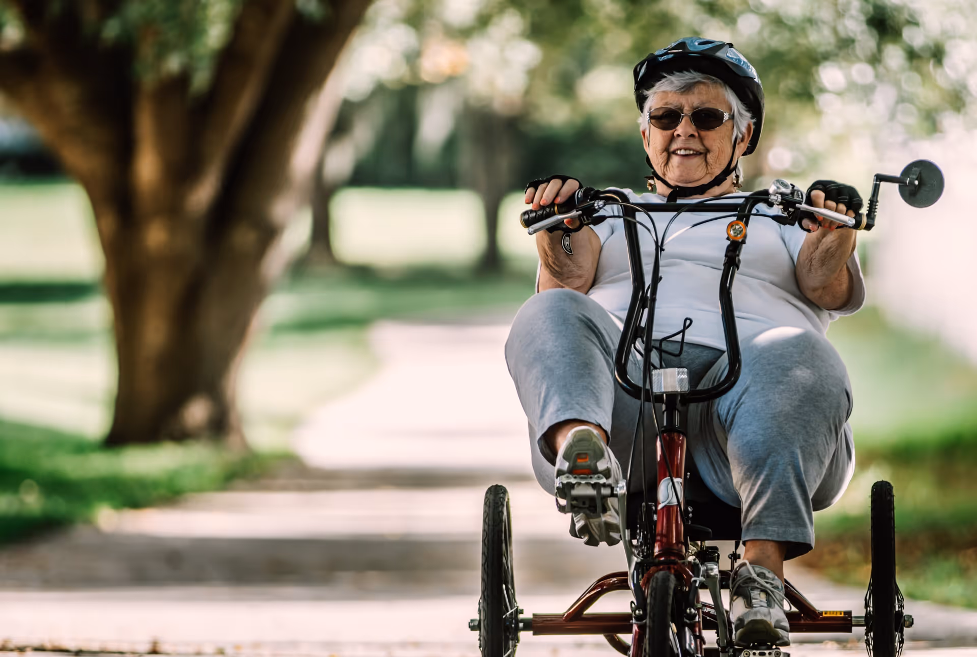 An elderly woman wearing a helmet and sunglasses rides a red three-wheeled recumbent bicycle on a paved path surrounded by green grass and trees.