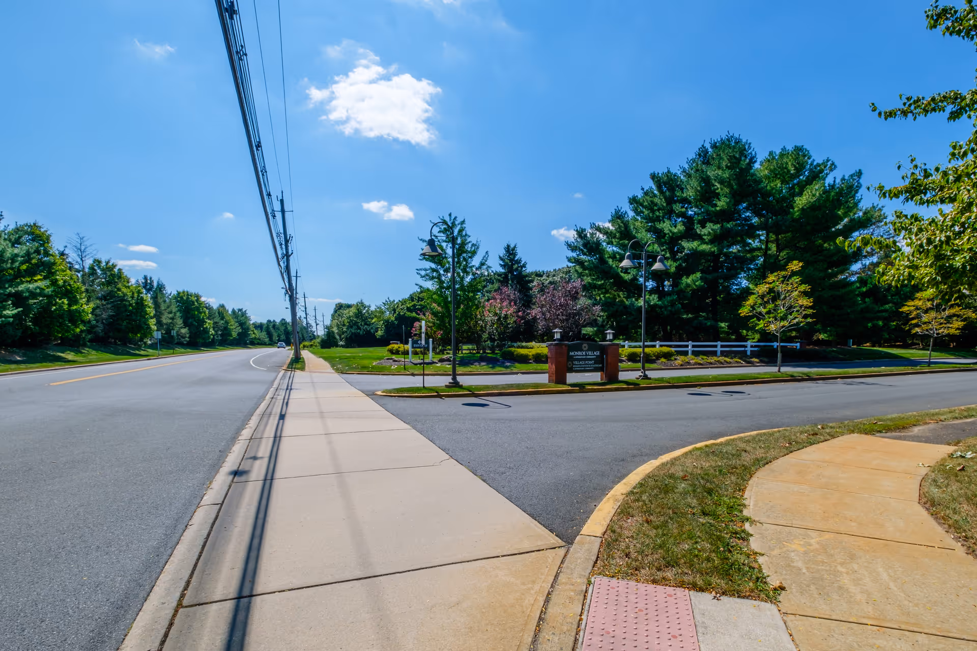 Sidewalk and roadway leading to a landscaped entrance sign for Monroe Village with trees and lamp posts under a blue sky.