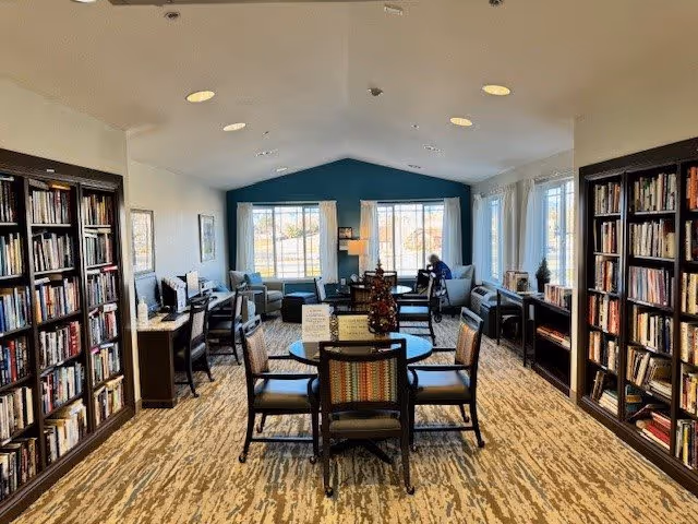 A bright and spacious library or reading room with bookshelves lining both sides of the room. There are tables and chairs in the center, with a few armchairs near large windows letting in natural light. A person is seated near the window on the right side, reading or working. The room has a carpeted floor and a high ceiling with recessed lighting.