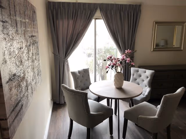 A small round dining table with a vase of pink flowers in the center, surrounded by four upholstered chairs with tufted backs. The room has wooden flooring, a large window with gray curtains, a dresser with a mirror above it, and a large abstract painting on the wall.