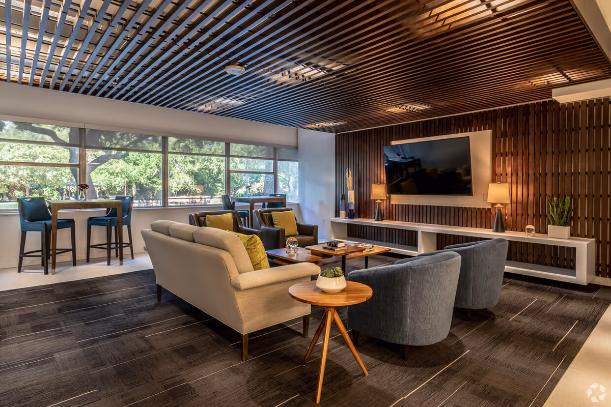 A modern senior living facility lounge area with a beige sofa, two blue armchairs, and two dark brown chairs around wooden coffee tables. The room features a large flat-screen TV mounted on a wooden slatted wall, two table lamps, and large windows showing greenery outside. The ceiling has wooden slats with recessed lighting, and there are two high tables with blue chairs near the windows.