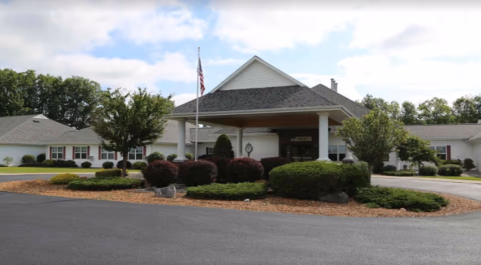 Exterior view of The Brook of Houghton Lake facility showing a single-story building with a covered entrance supported by white columns, surrounded by landscaped bushes and trees, with an American flag on a flagpole in front.