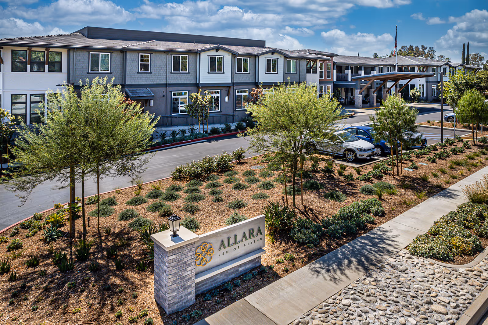 Exterior view of Allara Senior Living facility showing a two-story building with multiple windows, a landscaped garden with small trees and shrubs, a parking area with cars, and a sign with the facility's name at the front.