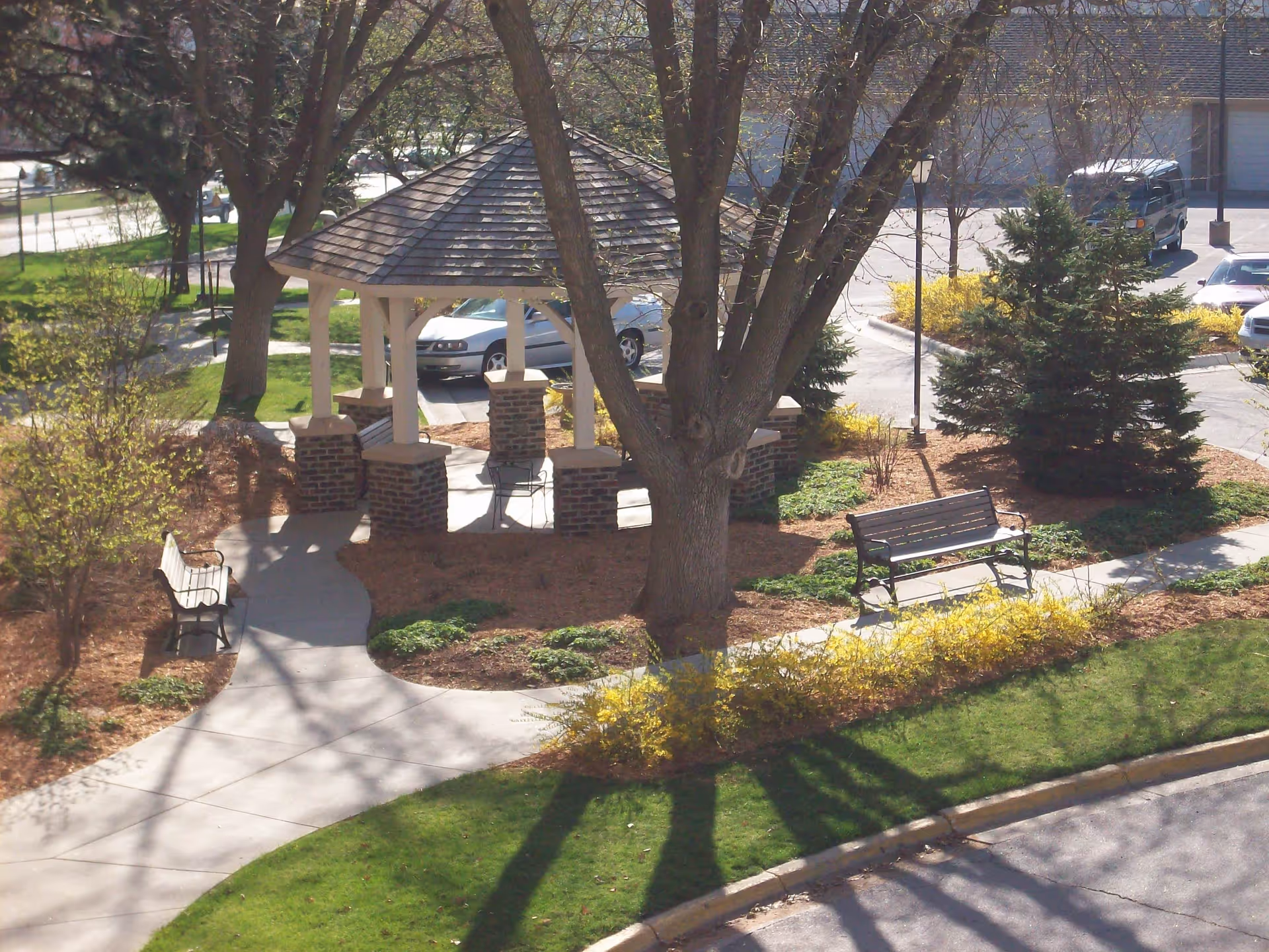 Outdoor garden area with a gazebo featuring a shingled roof and brick pillars, surrounded by trees, benches, and landscaped greenery. A parking lot with cars is visible in the background.