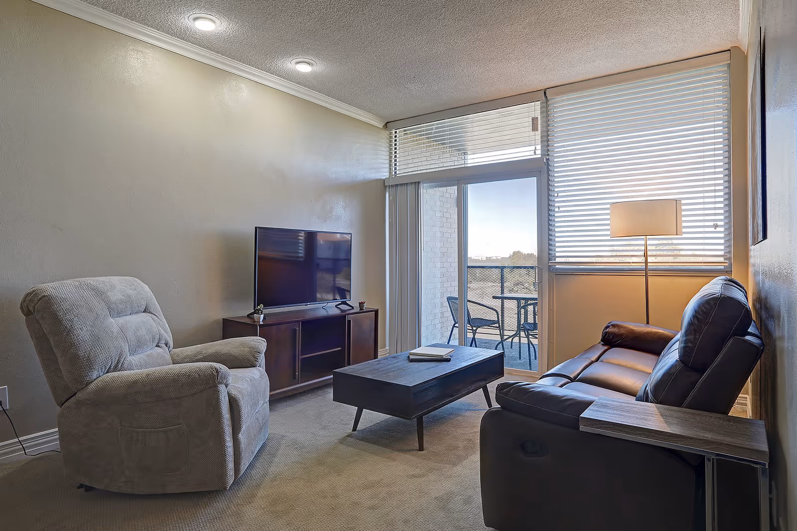 A cozy living room with a beige recliner chair, a dark brown leather sofa, a wooden coffee table with a book on it, a TV on a wooden stand, a floor lamp, and a sliding glass door leading to a balcony with outdoor chairs and a table. The room has beige walls and carpeted floor.