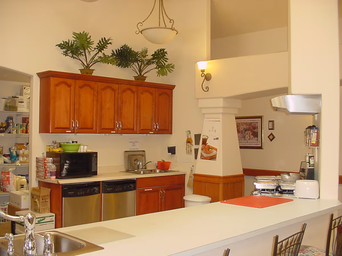 A kitchen area in an assisted living facility featuring wooden cabinets, a microwave, a dishwasher, a sink, and a countertop with bar stools. There are decorative plants on top of the cabinets and a pantry visible in the background.