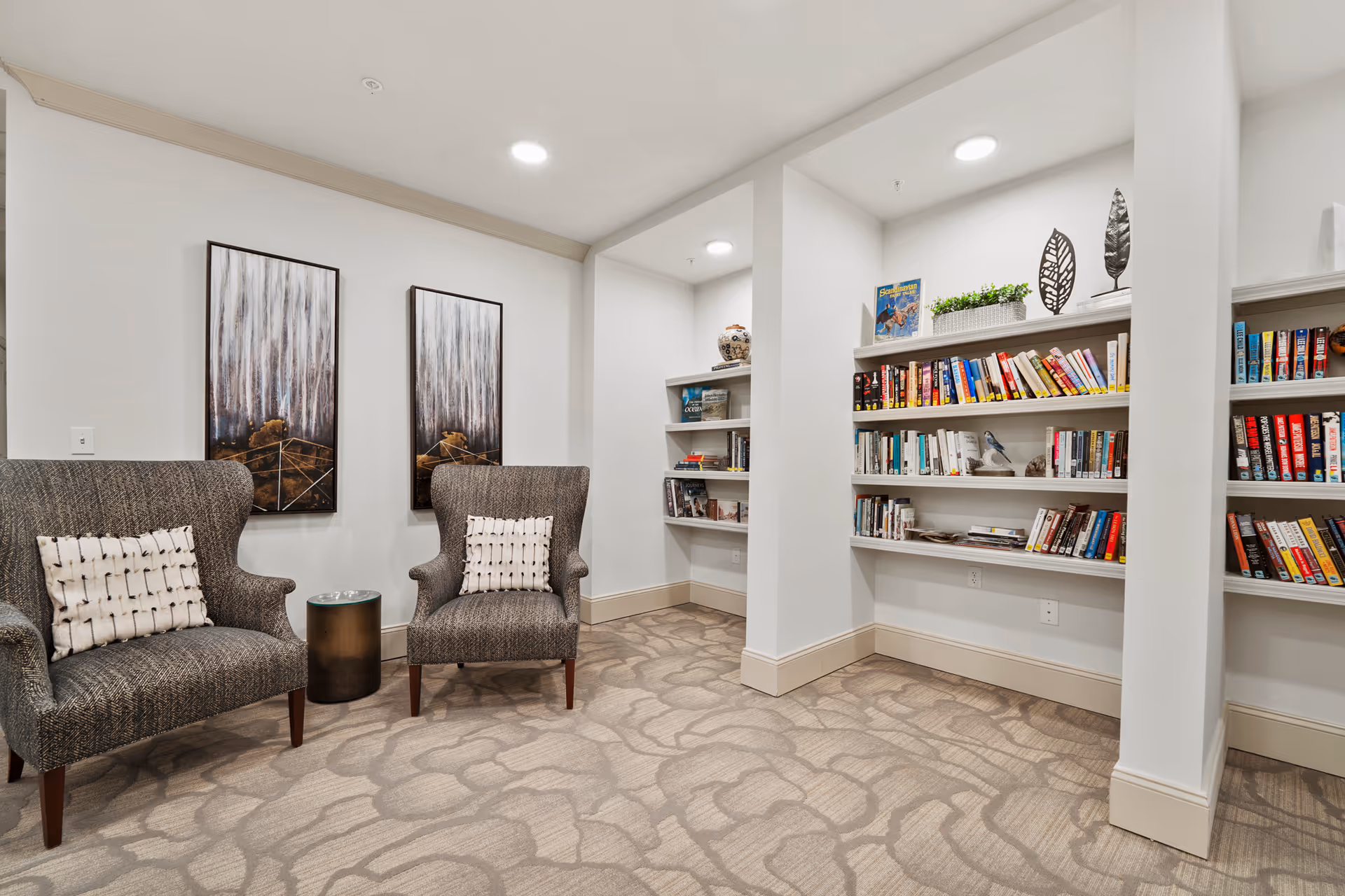 A cozy reading nook in a senior living facility featuring two upholstered armchairs with patterned cushions, a small round side table between them, and built-in white bookshelves filled with books and decorative items. The walls are white with two vertical abstract paintings, and the carpet has a subtle floral pattern.